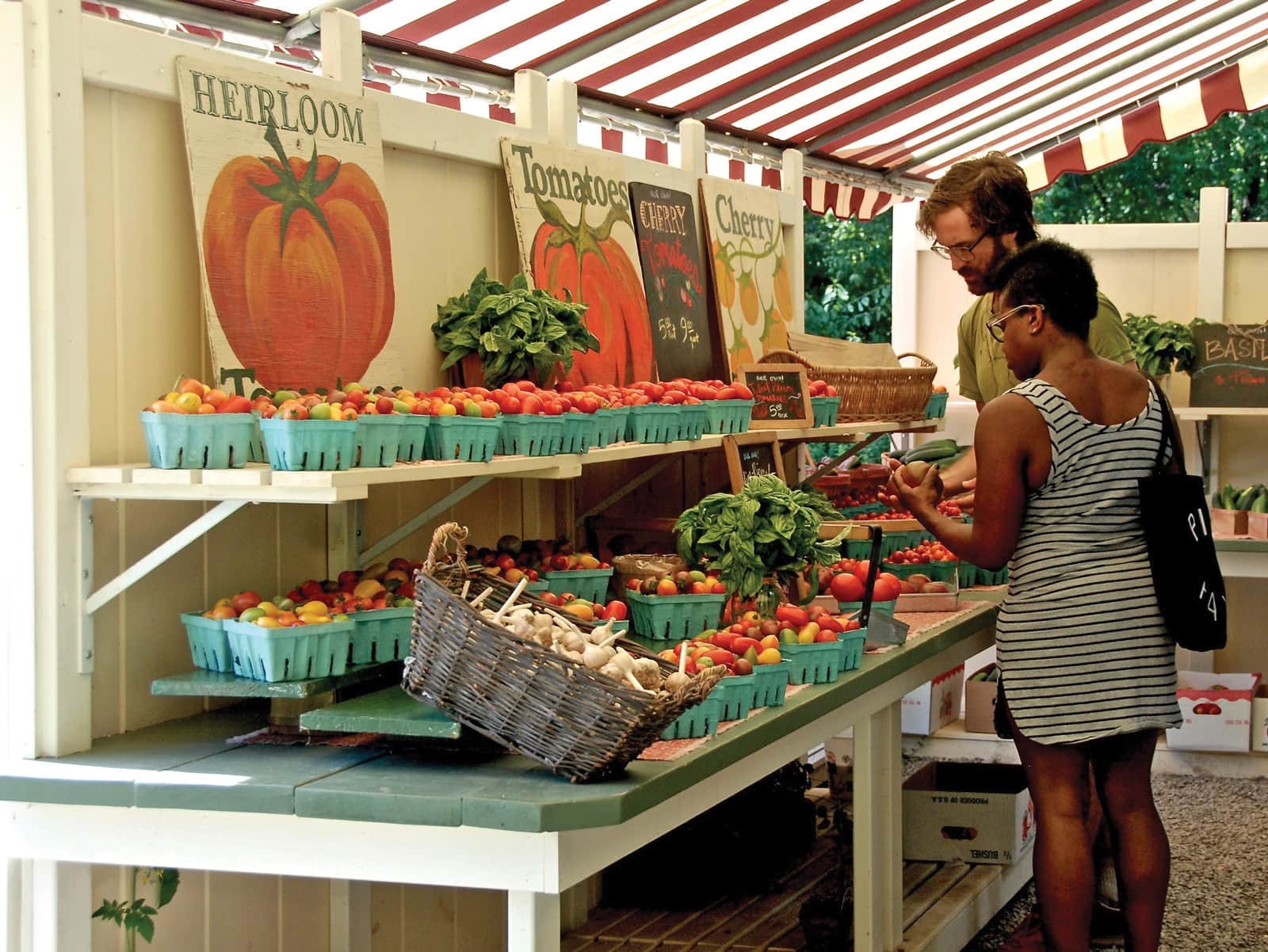 A welcoming roadside farm stand with hand-painted signs, baskets of heirloom tomatoes, and shoppers browsing under a striped awning